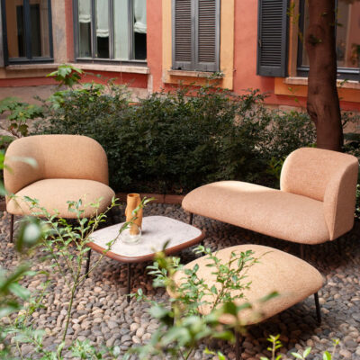 Outdoor seating area with peach-colored lounge chairs and a small marble-top table on a pebble ground, against a pastel building with shutters and greenery.