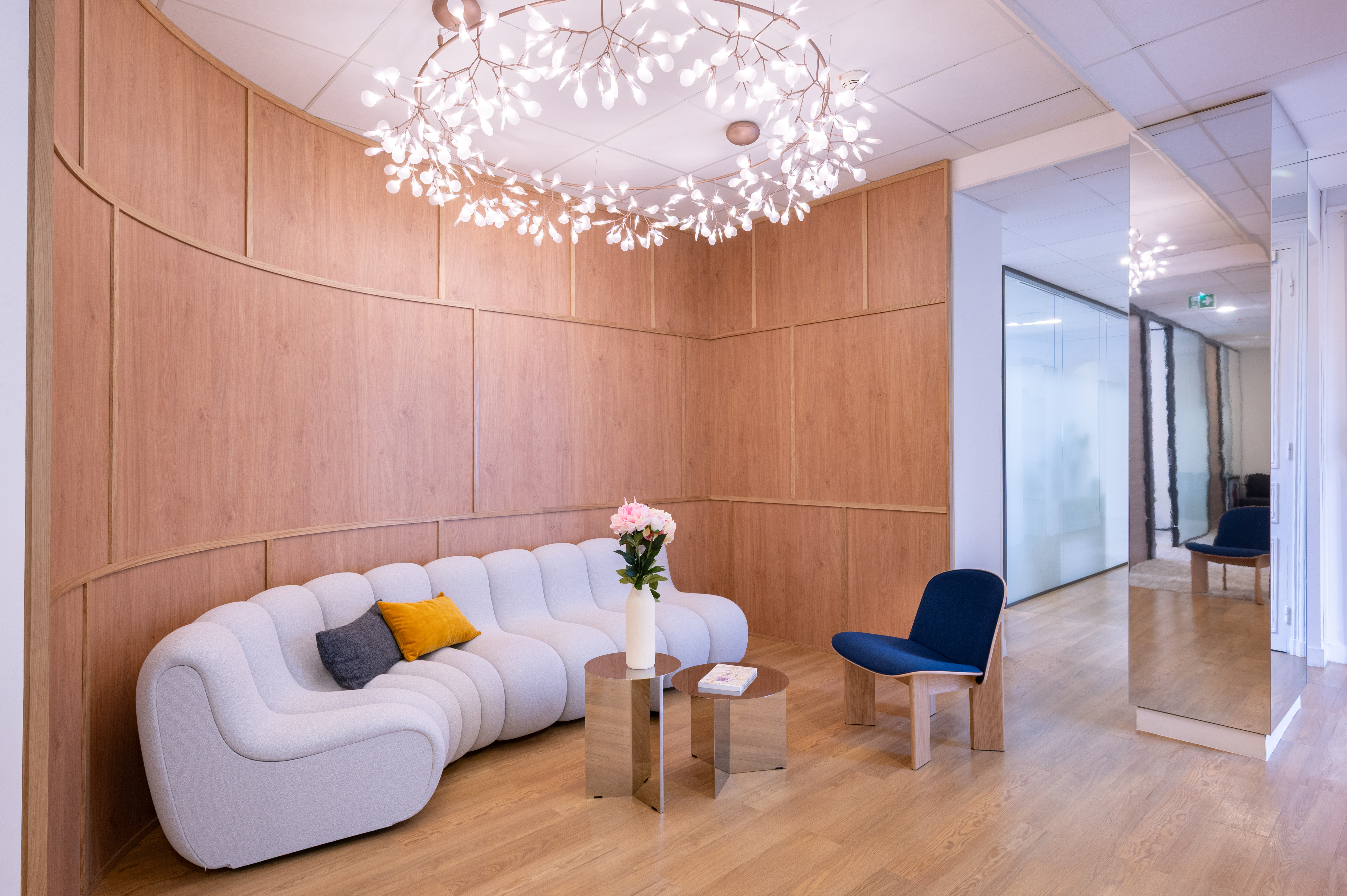 Curved wood-paneled wall with a white tufted sofa and small round tables in a modern lounge, a pink flower vase on the table and a branching chandelier overhead.