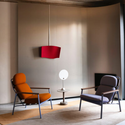Two mid‑century style armchairs (orange and purple) face each other with a small round table and white lampshade lamp between them in a minimal lounge setting, beige walls in the background.