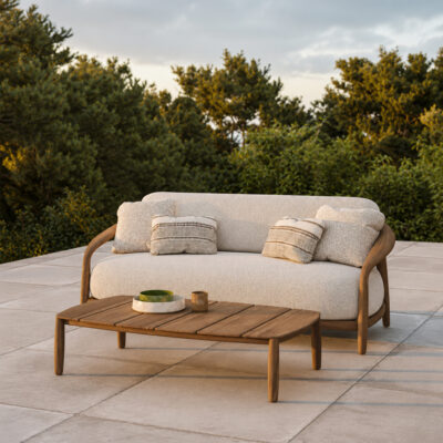 Outdoor patio seating: beige cushioned sofa with wooden frame and two patterned throw pillows, on a stone-tiled deck with greenery in the background.