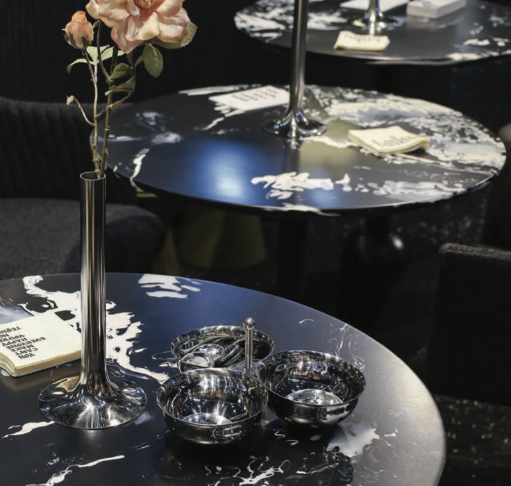 Round black marble-patterned table with a tall silver vase holding pale pink flowers and small matching silver bowls with tongs, in a chic lounge setting.