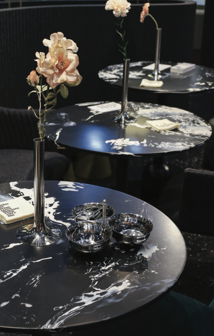 Round black marble-patterned table with a tall silver vase holding pale pink flowers and small matching silver bowls with tongs, in a chic lounge setting.
