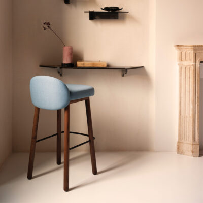 Blue upholstered bar stool with wooden legs against a beige wall, under a small wall shelf with a candle and book.