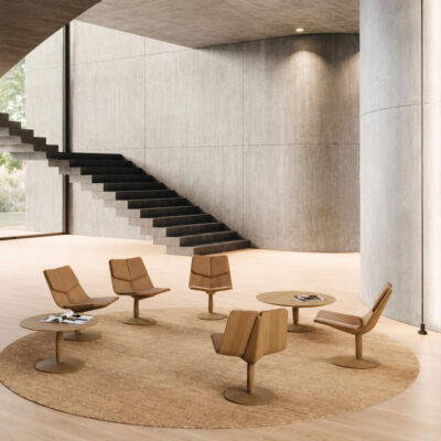 Six midcentury-style wooden chairs surround small round tables on a circular rug in a bright, concrete-walled lobby.