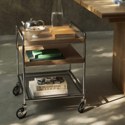 Rolling metal cart with two wooden shelves holding books and magazines; on top are a teapot, sugar bowl with cookies, and a glass bottle, with a yellowed book stack nearby; bottom shelf shows a vintage design book and a 'Food + Design' copy.