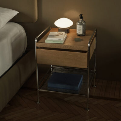 Wood and metal bedside nightstand with a rounded lamp, a bottle of lotion, magazines, and a lower shelf holding blue notebooks beside a bed.