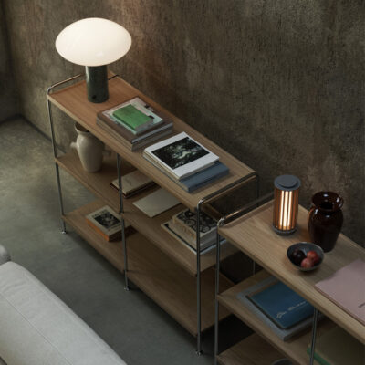 Three-tier wooden–metal shelf against a concrete wall with a white lamp, stacked books, vases, and a fruit bowl.