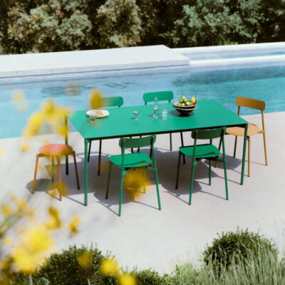 Green rectangular table with mixed-color chairs around it beside a pool, with a bowl of fruit and glassware on the table.